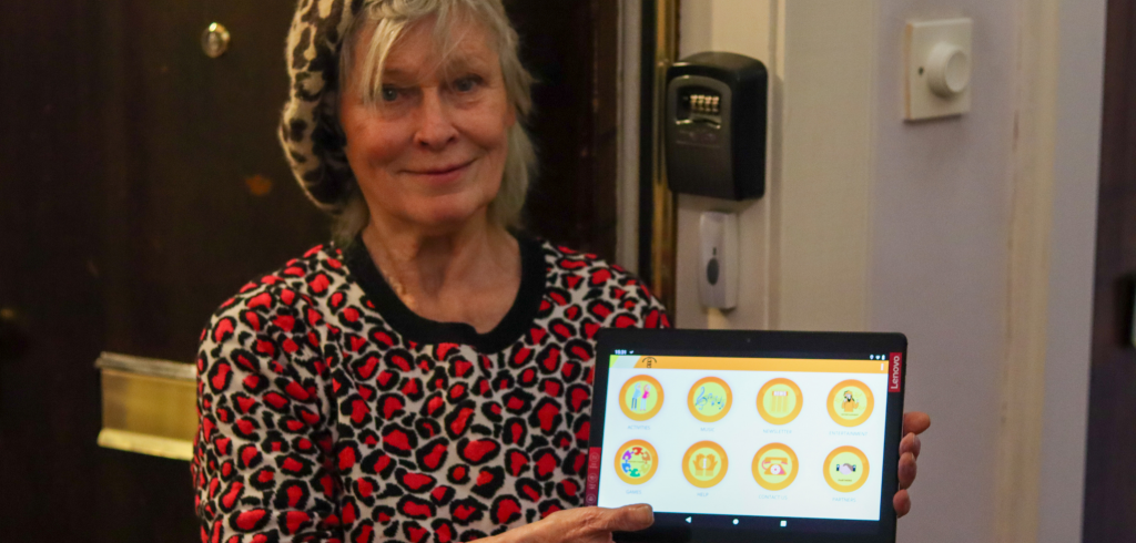 Picture of older adult in front of her door, holding a tablet with icons displayed